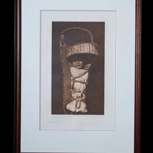 An Apache Babe photogravure by Edward S. Curtis, 1903 - Apache cradle board with infant wrapped in white fabric beneath a wooden sun shade