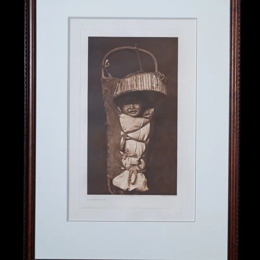 An Apache Babe photogravure by Edward S. Curtis, 1903 - Apache cradle board with infant wrapped in white fabric beneath a wooden sun shade